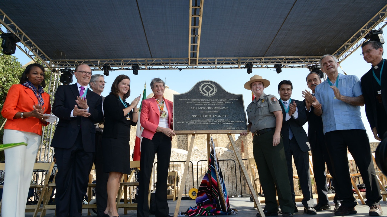 Cyclists riding along the Mission Reach trail with historic mission buildings in the distance.
