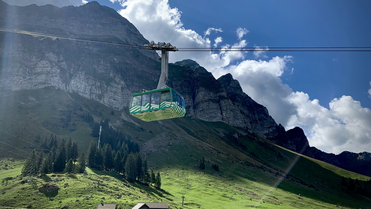 An aerial tram rising above a mountain ridge with passengers looking at panoramic views.