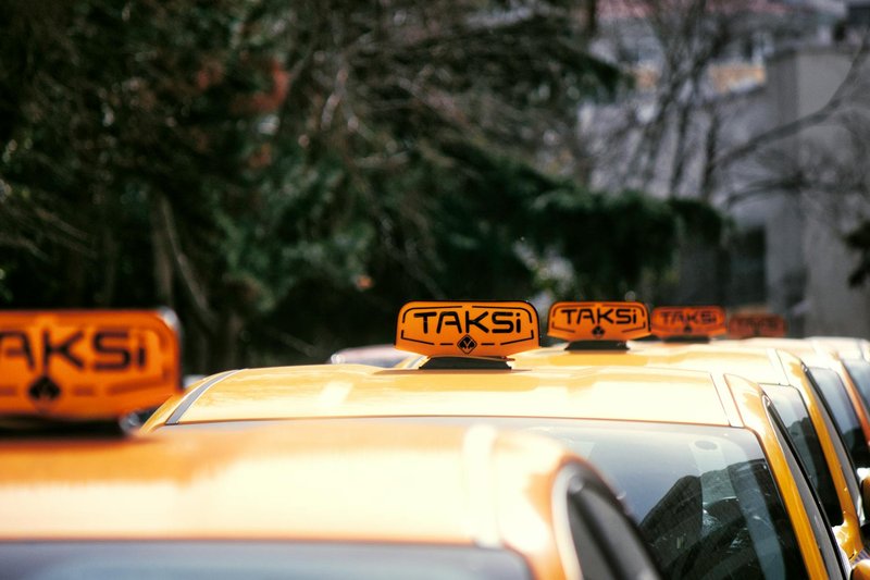 A row of yellow taxis with 'Taksi' signs parked on a city street in daylight.