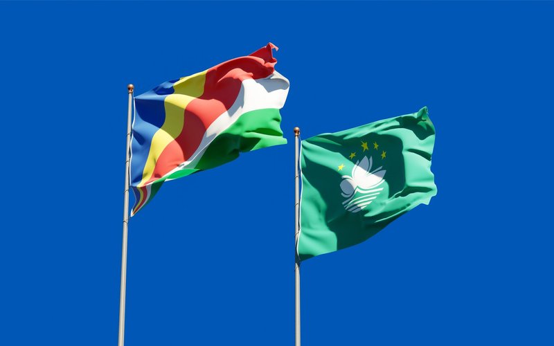 Seychelles and Macau flags waving on flagpoles against a bright blue sky, symbolizing international unity.