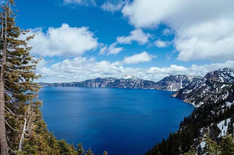 Stunning panorama of Crater Lake with clear blue waters and snow-capped mountains under a cloudy sky.