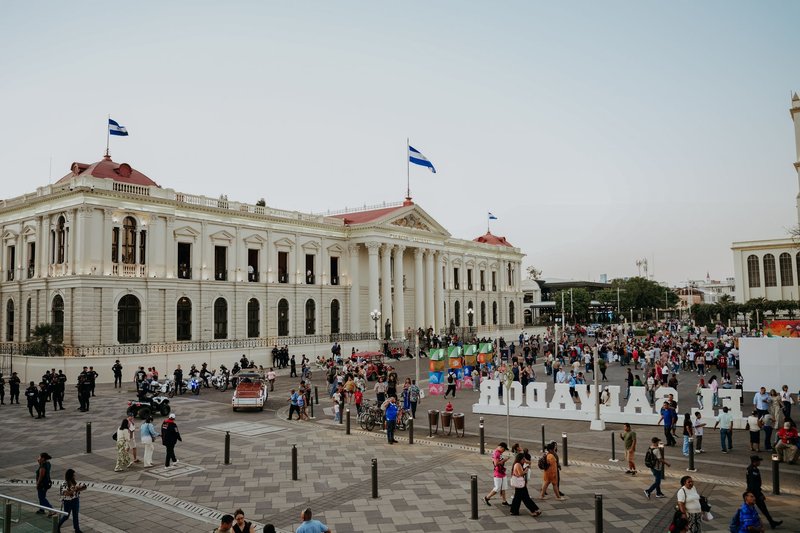 Crowded plaza scene in San Salvador with flags and historic buildings under a clear sky.