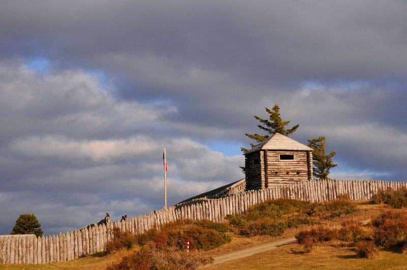 Rustic wooden fort surrounded by fencing on a grassy hill under a dramatic cloudy sky.