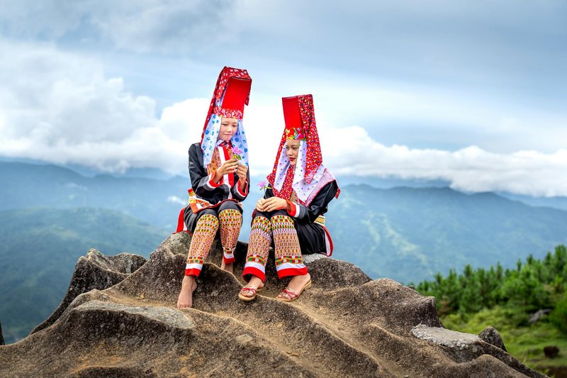 Two women in traditional attire pose on a mountain with clouds in the background.