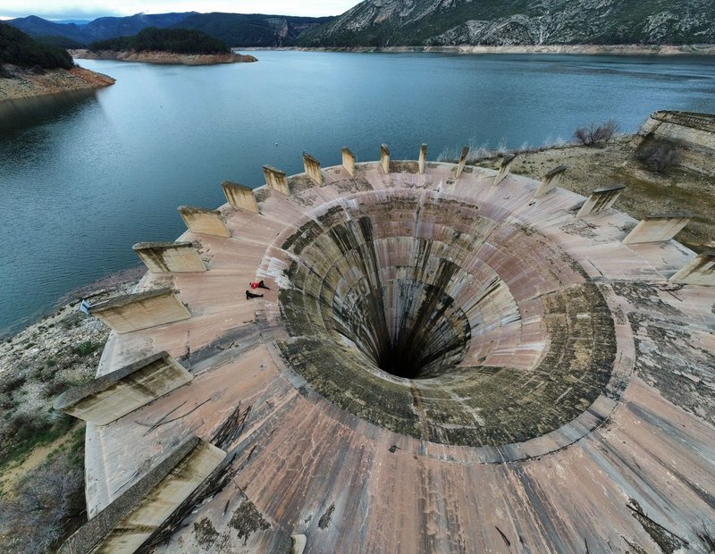 Aerial view of a large drainage spillway at a scenic reservoir surrounded by hills.