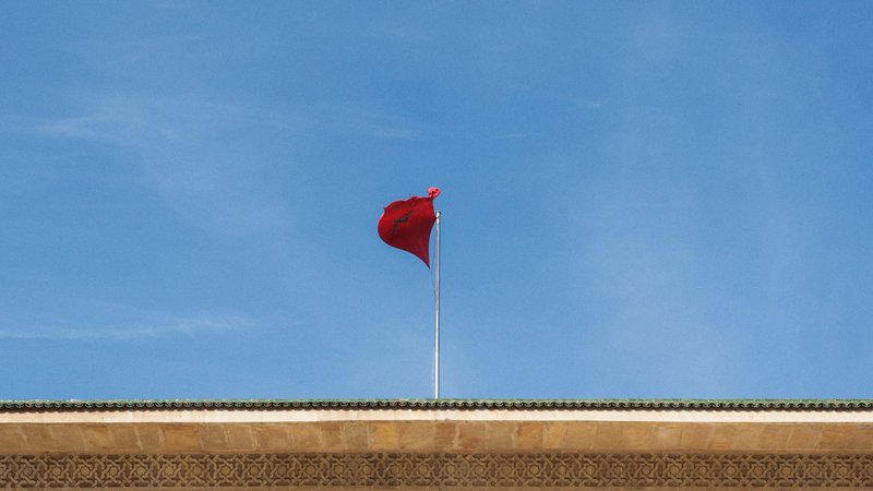 Red Moroccan flag atop building with ornate patterns against clear blue sky.