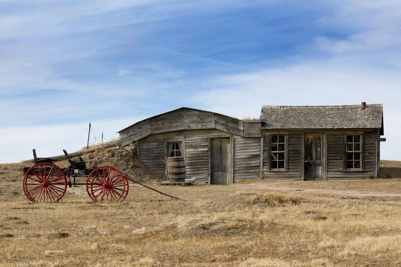 Rustic wooden farmhouse with a vintage carriage on a grassy prairie.