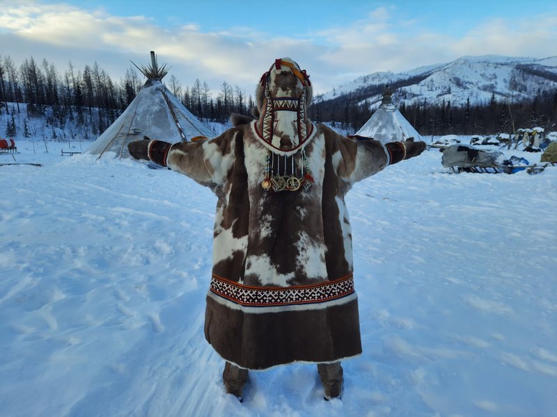 Person in traditional Siberian attire in a snowy landscape with tents and trees.