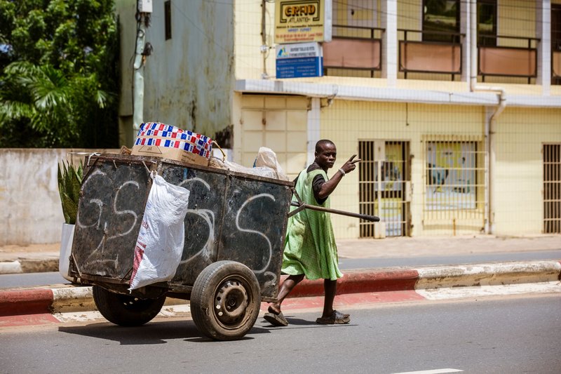 A street vendor in a green dress pushes a cart along an urban street in Lomé, Togo.