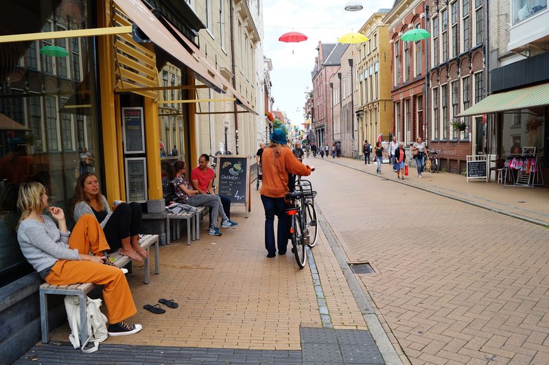 Urban street scene with people walking and biking, vibrant shops and cafes on a sunny day.