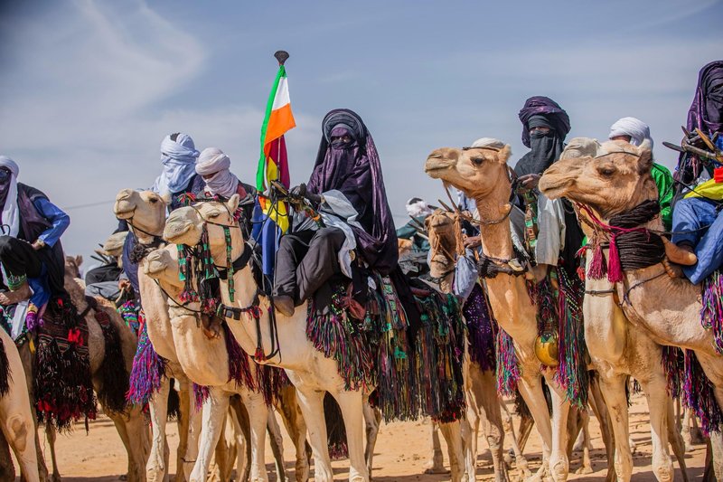 Tuareg men in traditional attire on camels with Niger flag in desert parade.