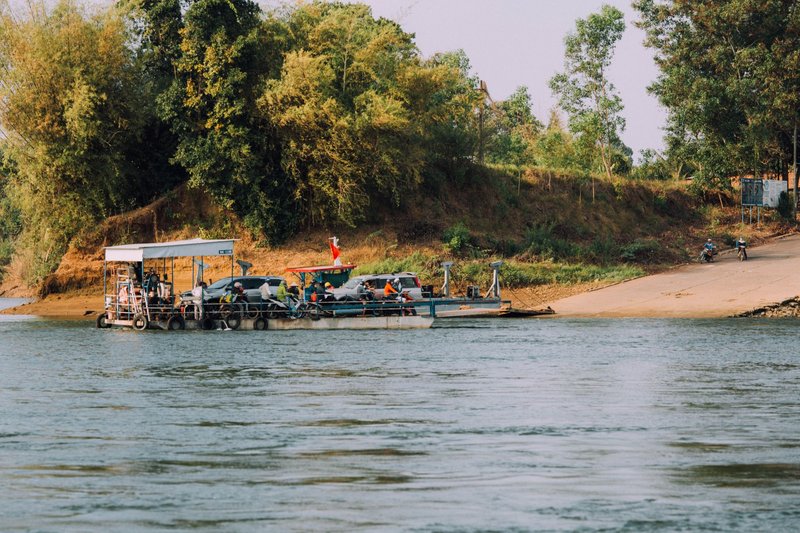 A vibrant river ferry transporting vehicles and passengers, set against a lush green backdrop.