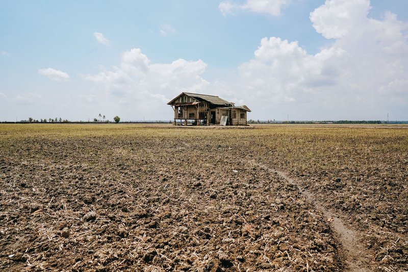 A lonely abandoned farmhouse sits in a wide, empty field under a bright sky.
