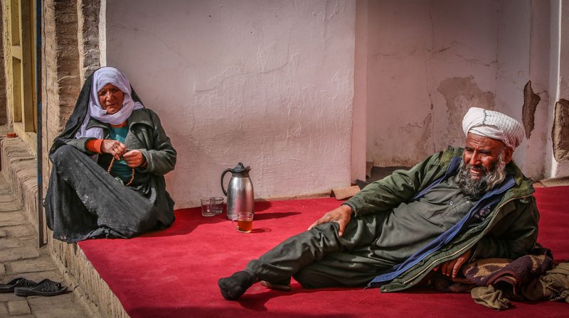 An elderly Afghan couple resting on a vibrant red carpet in Herat, Afghanistan, showcasing traditional attire and daily life.