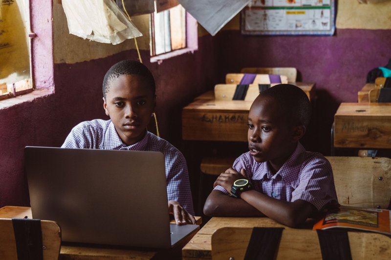 Two black boys in school uniforms focusing on a laptop, collaborating in a classroom setting.