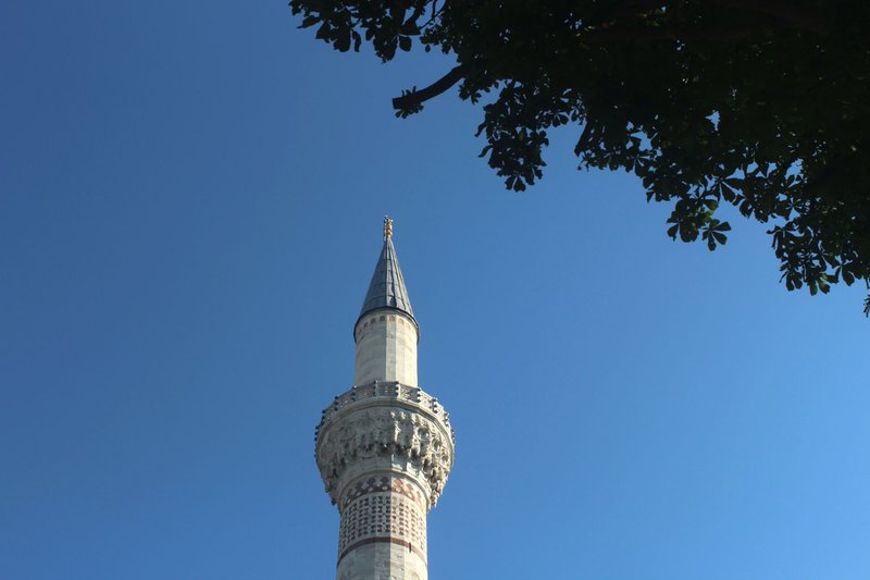 A detailed view of a minaret with ornate design under a clear blue sky, framed by tree foliage.