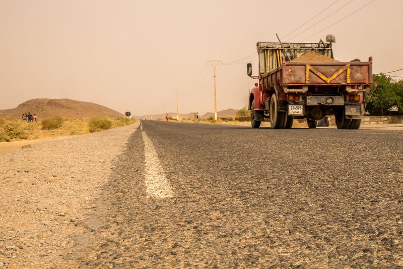 A red pickup truck drives on a long desert road under a hazy sky.