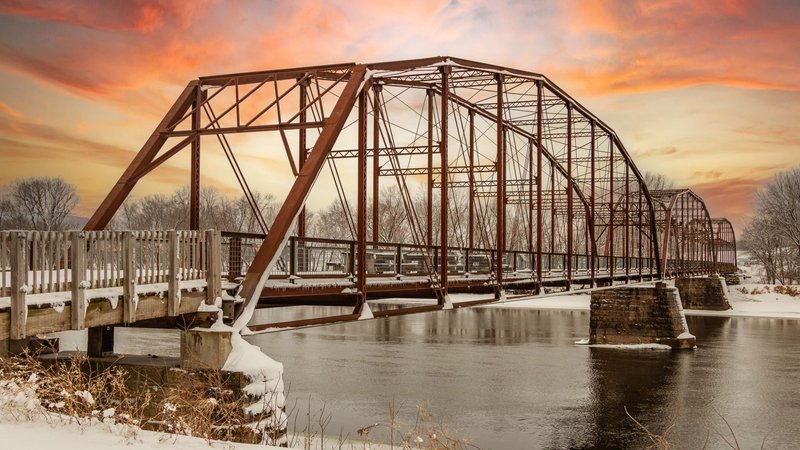A scenic view of a historic steel truss bridge over a river during a colorful winter sunset.