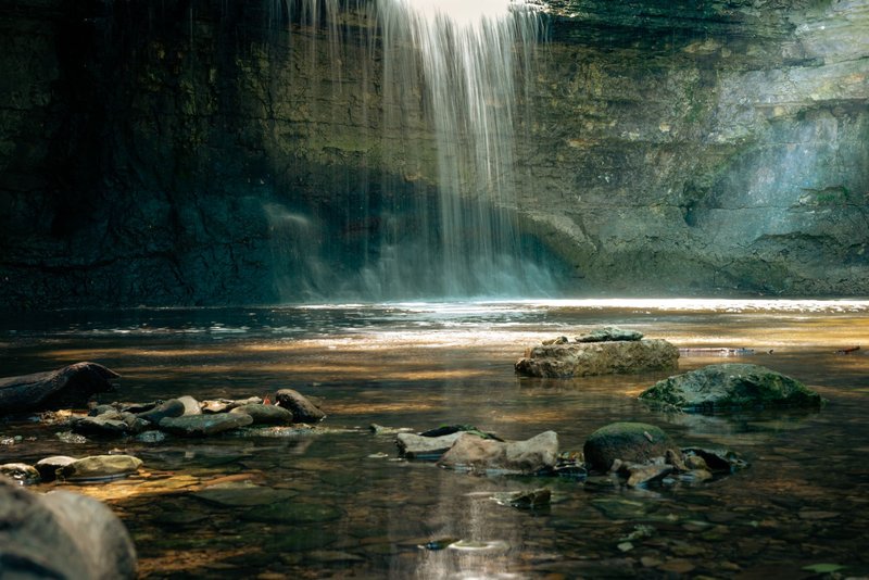 Beautiful waterfall cascading into a serene pool, surrounded by rocks, under a canopy of sunlight.