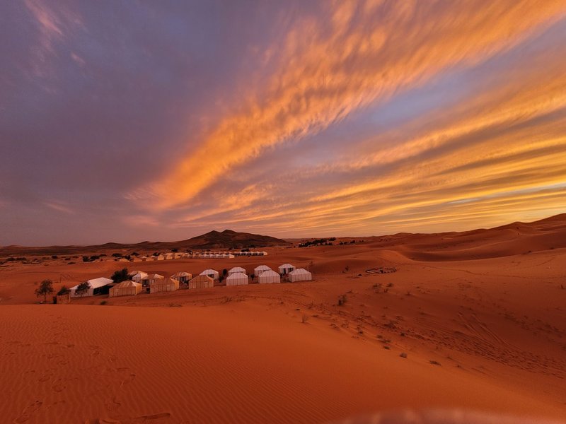 White tents in the Sahara Desert under a dramatic orange and blue sunset sky.