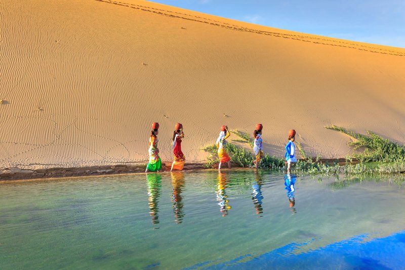 Colorfully dressed women walking by a desert oasis with water jars, creating vibrant reflections.