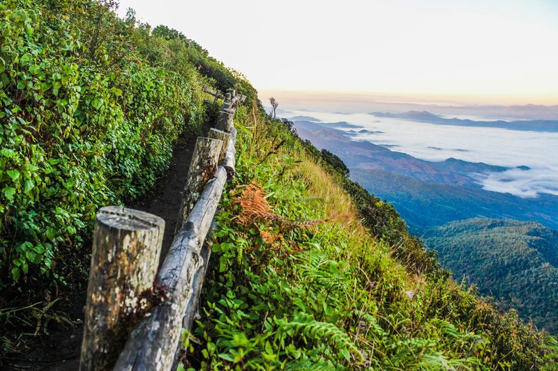 Scenic mountain path with wooden fence overlooking a verdant valley and clouds.