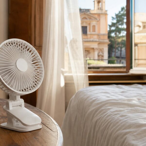 White clip-on travel fan on a wooden nightstand in a sunlit European hotel room with open shutters