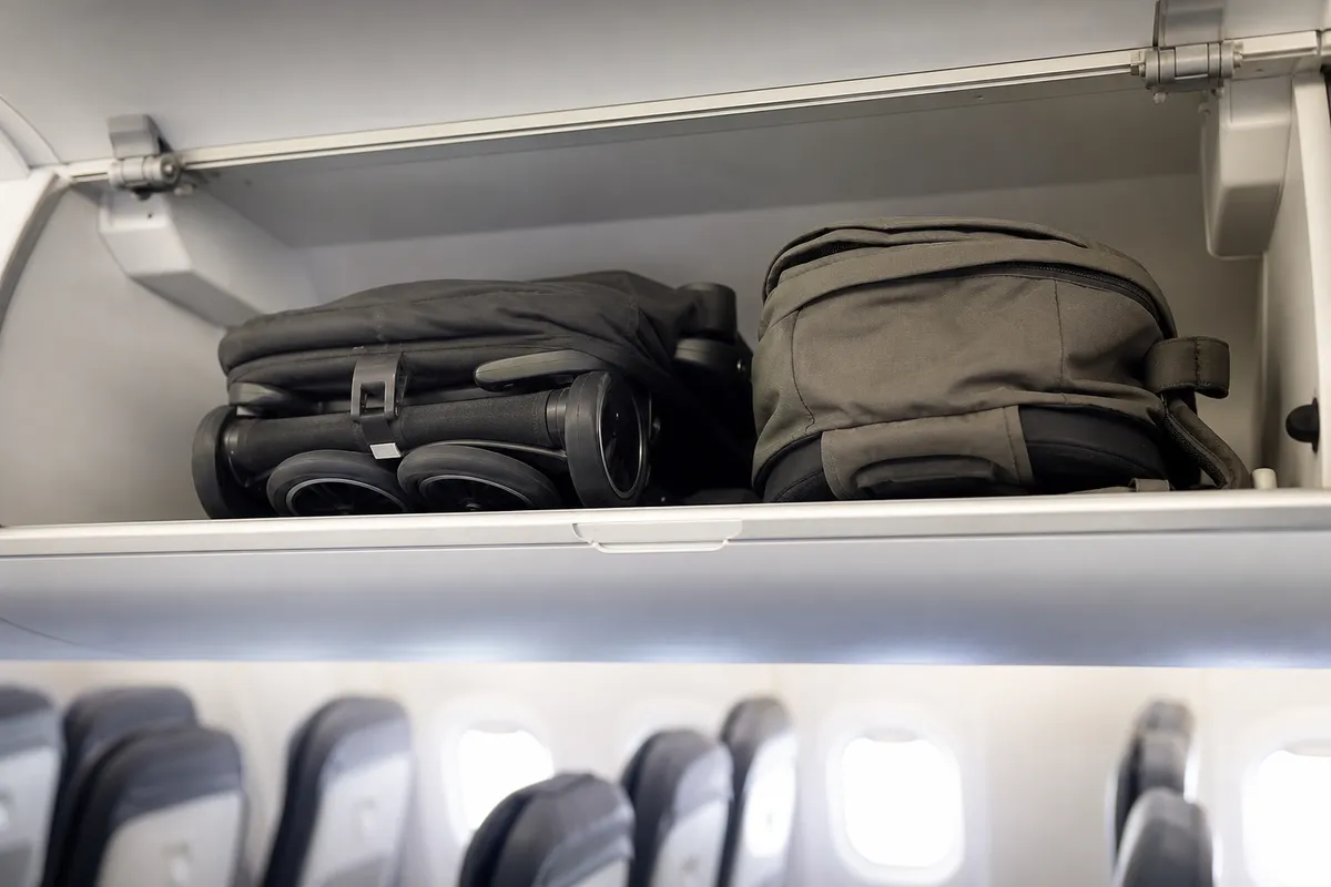 Folded compact stroller lying in an open airplane overhead bin next to a backpack