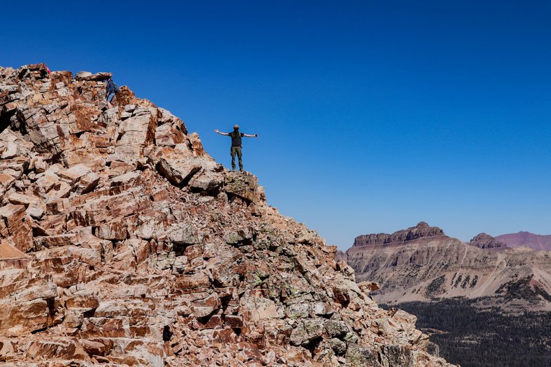 A climber celebrates atop a rugged mountain peak under a clear blue sky.