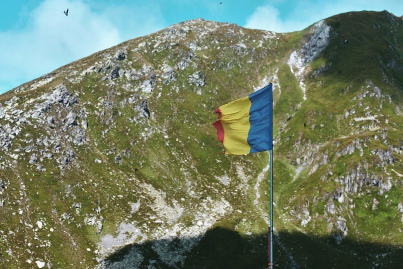 Romanian flag waves atop a scenic mountain landscape under a clear blue sky.