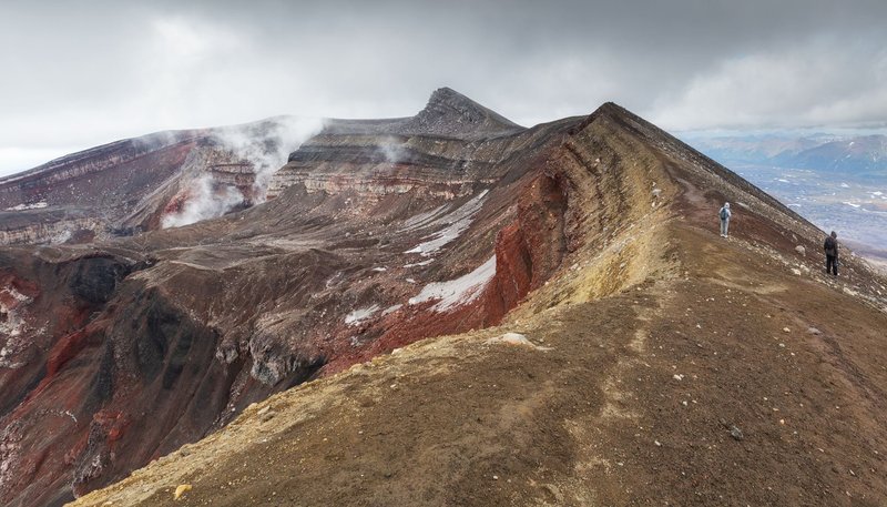 Hikers explore a vibrant volcanic ridge with breathtaking mountain views under cloudy skies.
