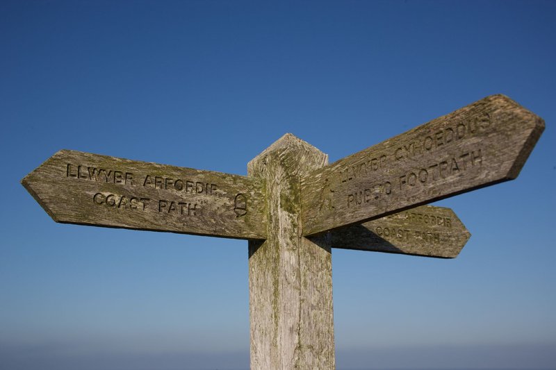A rustic wooden signpost on the Welsh Coastal Path under a clear blue sky.