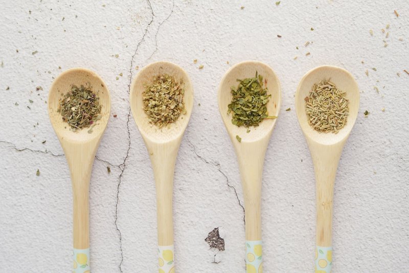 Four wooden spoons with assorted dried herbs on a light background.