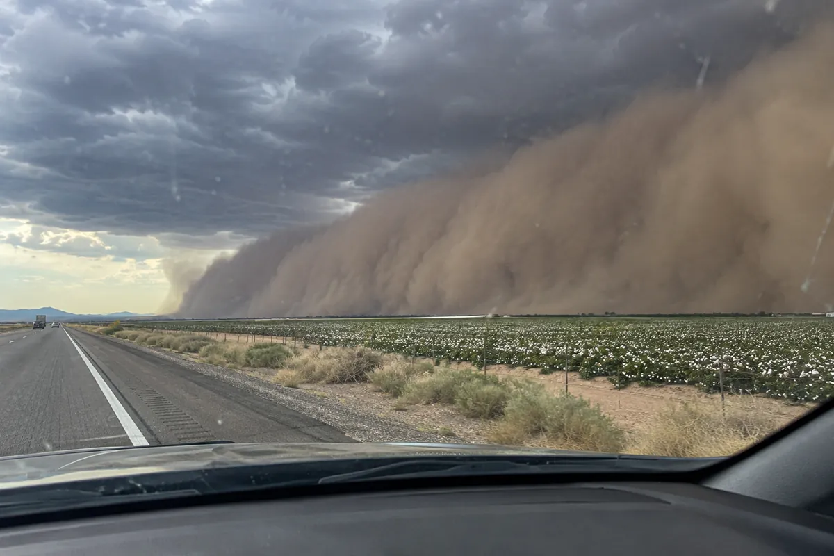 A brown dust storm rolling over farmland toward a desert interstate under dark clouds