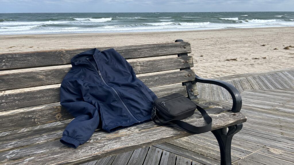 Navy windbreaker and black crossbody bag on a wooden bench on the Atlantic City Boardwalk, ocean behind