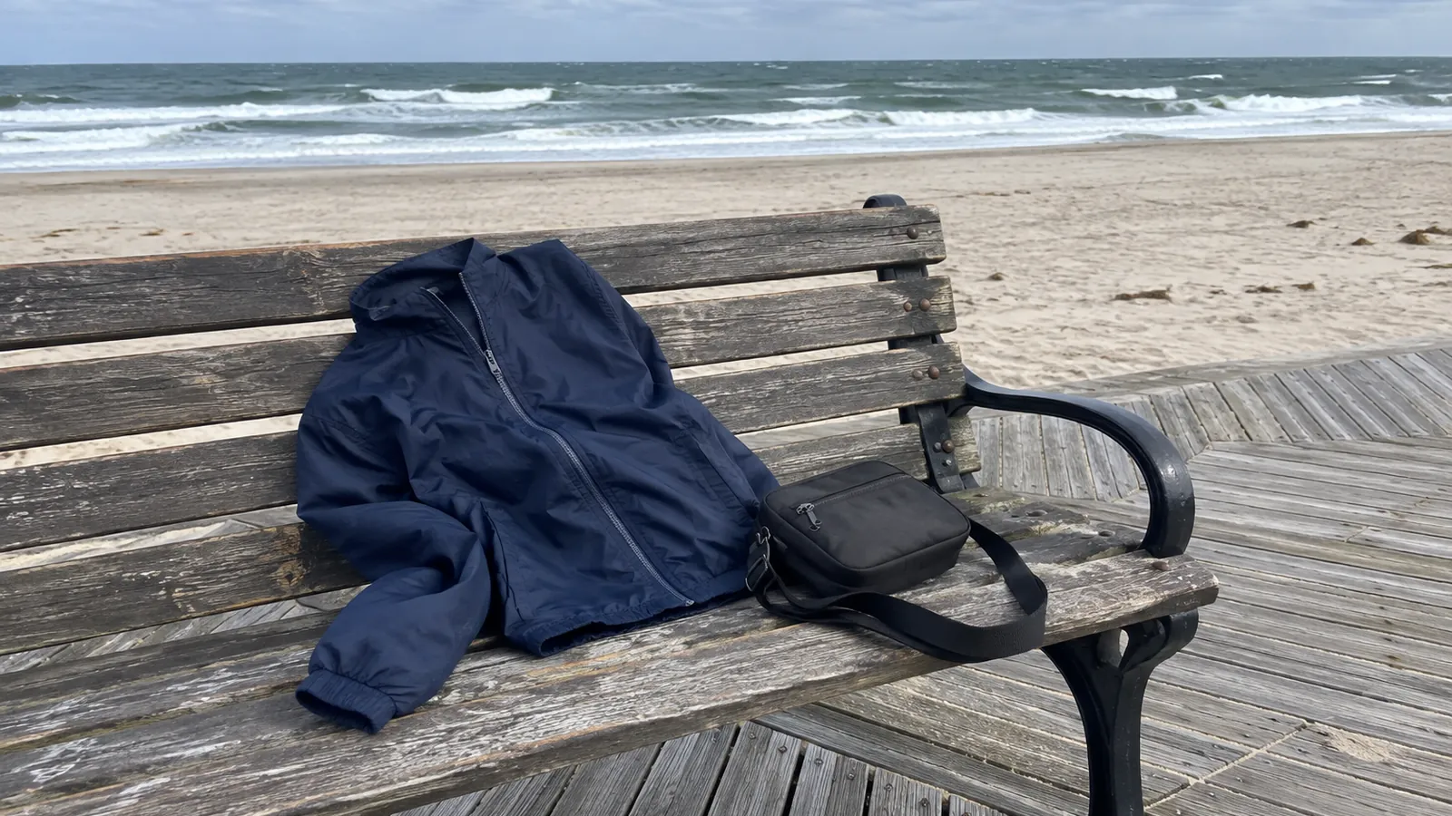 Navy windbreaker and black crossbody bag on a wooden bench on the Atlantic City Boardwalk, ocean behind