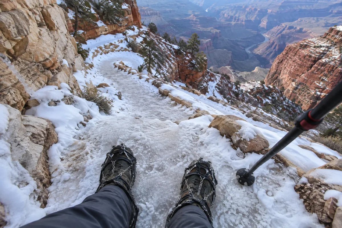 Feet in trail shoes with chain traction on packed icy snow on a switchback below the canyon rim, a trekking pole in frame