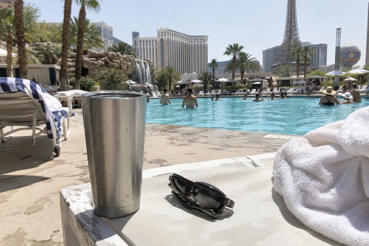 Insulated steel tumbler and sunglasses on a white lounge chair beside a resort pool under harsh June sun