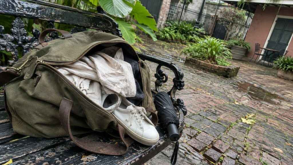 Open canvas weekender with a linen shirt and white sneakers on an iron bench in a wet brick French Quarter courtyard