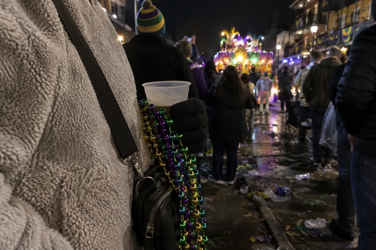 A gloved hand holding purple, green and gold beads and a plastic cup on a crowded parade route at night