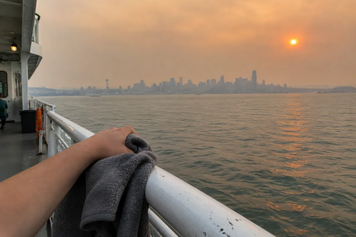 Forearm resting on a ferry rail holding a folded fleece, hazy orange sky and a faint skyline across the water